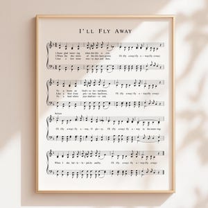 May include: A framed print featuring sheet music with the title "I'll Fly Away." The musical notes and lyrics are printed in black on a cream-colored background. The frame is a thin, gold color, adding a touch of elegance to the artwork.