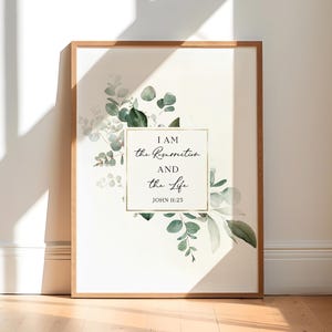 May include: A framed print with a white background and a gold frame. The print features a watercolor illustration of green eucalyptus leaves and the text "I AM the Resurrection and the Life JOHN 11:25".