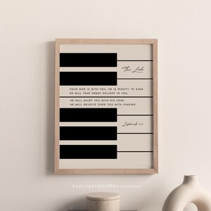 Op de afbeelding: Ingelijste kunstdruk met een pianotoetsontwerp, zwarte toetsen en een beige achtergrond. De tekst bevat een schriftgedeelte uit Zefanja 3:17. De lijst heeft een lichte houtkleur.