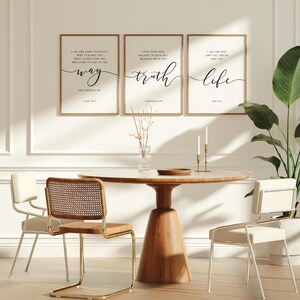 May include: A dining room setting with a round wooden table and three framed prints with inspirational text. The prints are above the table, which is surrounded by three chairs. A vase with dried flowers and two candlesticks are on the table.