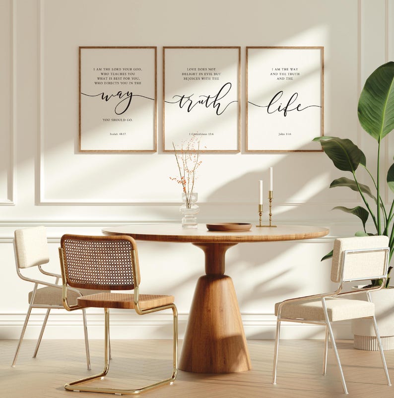 May include: A dining room setting with a round wooden table and three framed prints with inspirational text. The prints are above the table, which is surrounded by three chairs. A vase with dried flowers and two candlesticks are on the table.