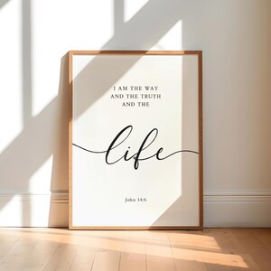 May include: A framed print featuring the words "I AM THE WAY AND THE TRUTH AND THE LIFE" in black script. The print is in a light wood frame and leans against a white wall. The text "John 14:6" is at the bottom.