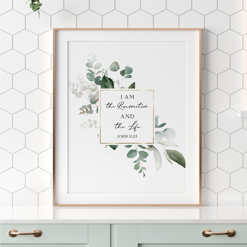 Puede incluir: Una impresi&oacute;n enmarcada en blanco con un borde dorado que presenta una ilustraci&oacute;n de acuarela de hojas de eucalipto verdes y flores blancas. El texto "I AM the Resurrection AND the Life JOHN 11:25" est&aacute; escrito en letra cursiva negra.