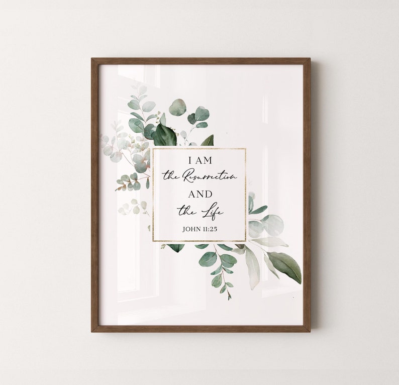 Puede incluir: Una impresi&oacute;n enmarcada con fondo blanco y marco dorado. La impresi&oacute;n presenta una ilustraci&oacute;n de acuarela de hojas de eucalipto verdes y flores blancas. El texto "I AM the Resurrection and the Life JOHN 11:25" est&aacute; impreso en letra cursiva negra dentro de un rect&aacute;ngulo dorado.