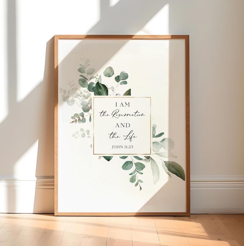 May include: A framed print with a white background and a gold frame. The print features a watercolor illustration of green eucalyptus leaves and the text "I AM the Resurrection and the Life JOHN 11:25".