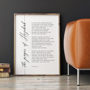 May include: A framed print with the text "The prayer of Hezekiah" in elegant script. The print features a passage from 2 Kings 20:1-5, set against a white background. The frame is a light wood color, and the print is displayed near a leather chair.