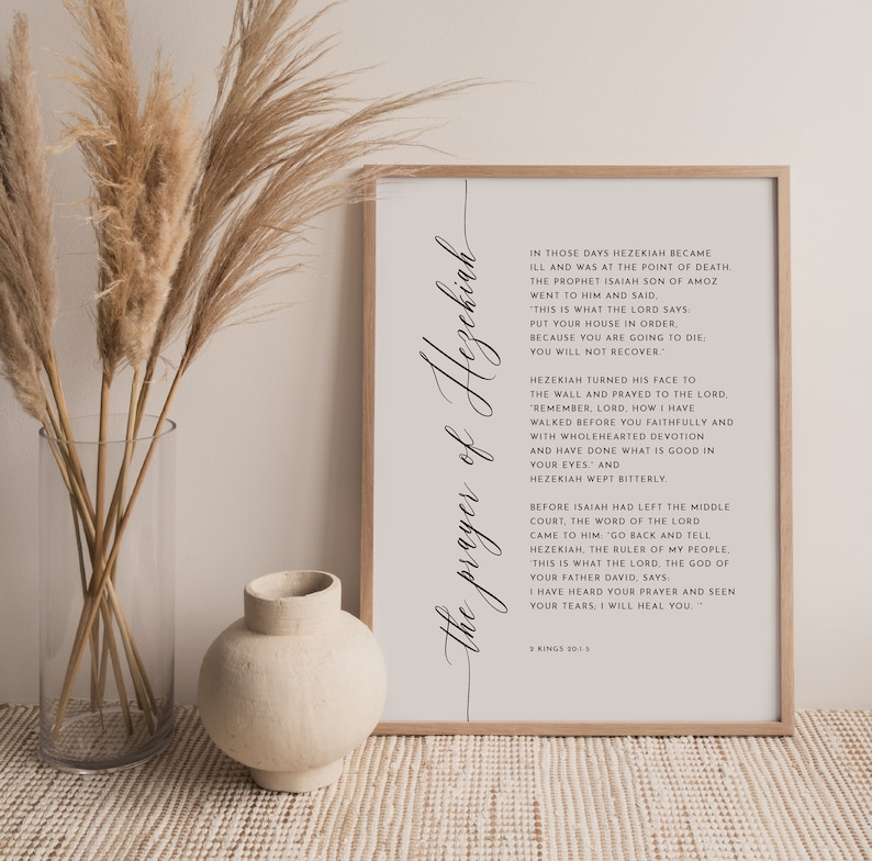 May include: A framed print with the text "the prayer of Hezekiah" in elegant script. The print features a beige background with black text from 2 Kings 20:1-5. The frame is a light wood color, and the image is displayed with dried plants and a vase.