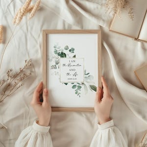 Puede incluir: Una impresi&oacute;n enmarcada con una ilustraci&oacute;n de acuarela de hojas de eucalipto y flores blancas. La impresi&oacute;n presenta el texto "YO SOY la resurrecci&oacute;n y la vida" con la referencia b&iacute;blica "JUAN 11:25".