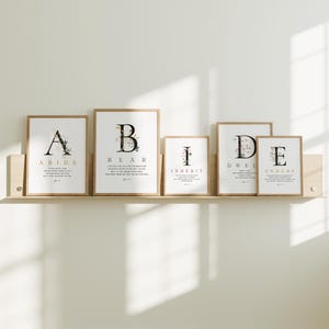 May include: Set of six framed prints with floral designs and gold lettering. Each print features a different letter of the alphabet and a corresponding word: ABIDE, BEAR, INHERIT, DWEL, ENDURE. The prints are arranged in a row on a wooden shelf.