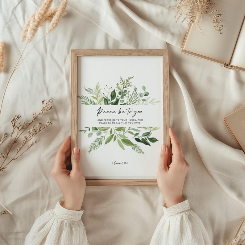 May include: A framed print with the text "Peace be to you." and botanical illustrations in green. The print is held by hands and has a light wood frame. The background is a soft, neutral fabric.