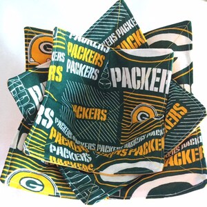 May include: A stack of green and yellow fabric bowl cozies. The cozies are patterned with the words "PACKERS" and the team's logo. The fabric has a quilted design. The cozies are square and have a pocket on one side.