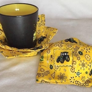 May include: A black ceramic mug with a yellow interior sits in a yellow fabric bowl-shaped coaster. A second coaster is next to it. Both coasters feature a yellow bandana print with green tractors and floral designs.