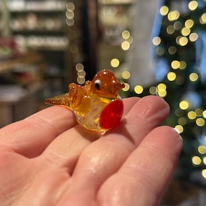 May include: A small, glass bird figurine with a red breast, yellow body, and brown head. The bird is perched on a hand, with a blurred background of lights and greenery. The figurine is likely a decorative item or ornament.