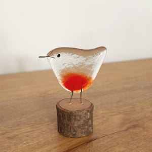 Op de afbeelding: Een vogelbeeldje van gefuseerd glas met een doorschijnend lichaam en een felrode borst. De vogel heeft een klein zwart oog, een dunne draad als snavel en draadpoten die vastzitten op een cilindervormige basis van natuurlijk schorshout.