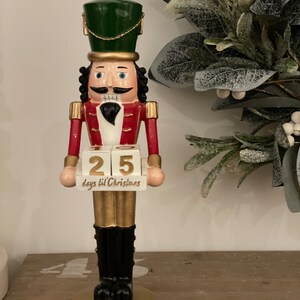 Può includere: Figurina decorativa schiaccianoci natalizia con cappello verde, giacca rossa e stivali neri. La figurina mostra "25 giorni a Natale". Dettagli e base dorati completano il design.