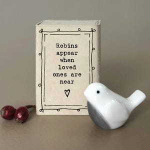 May include: A white ceramic bird ornament with a grey base is positioned beside a small box. The box features the text "Robins appear when loved ones are near" within a black frame on a cream background. Two red berries are in the foreground.
