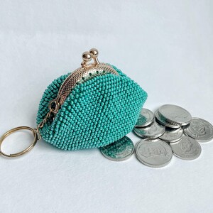 May include: A teal beaded coin purse with a gold-colored clasp and key ring. Several silver coins are scattered beside the purse. The purse is covered in small, round beads, and the clasp has a decorative design.