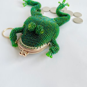 以下が含まれることがあります： 金色の留め具と2つの金色のノブが付いた緑色のビーズでできたカエルの財布。財布は白い表面に置かれ、その周りにいくつかの銀貨が散らばっている。