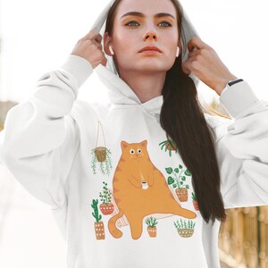 Peut inclure: Sweat-shirt blanc avec un motif d'un chat orange assis parmi des plantes en pot. Le chat tient une tasse de café.