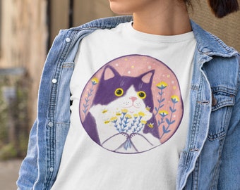 Puede incluir: Camiseta blanca con un gráfico de un gato blanco y negro sosteniendo un ramo de flores moradas con abejas amarillas. El gato está rodeado de un círculo rosa con un fondo rosa claro.
