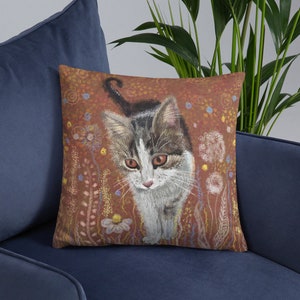 May include: A throw pillow with a colorful illustration of a tabby cat walking through a field of flowers. The cat has white fur with brown and gray stripes. The background is a warm brown color with a variety of flowers and plants in shades of pink, purple, and yellow.