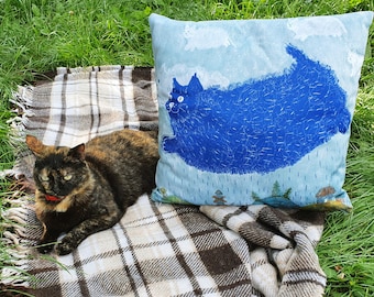 May include: A brown and white plaid blanket with a blue cat pillow featuring a blue cat with a white face and large eyes. A brown and black cat is lying on the blanket.