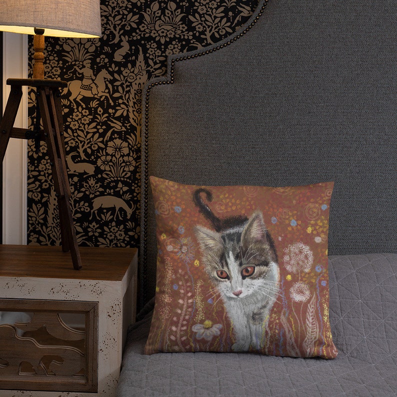 May include: A throw pillow with a brown background featuring a white and gray kitten walking through a field of flowers. The flowers are in shades of pink, purple, and yellow.