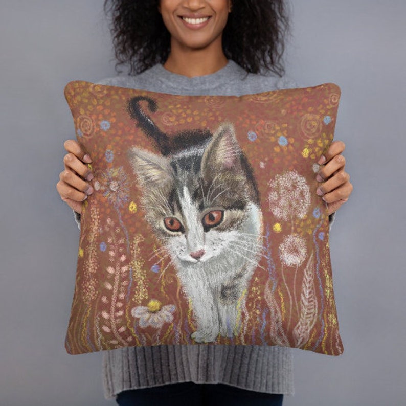 May include: A brown and orange throw pillow with a hand-painted illustration of a white and gray cat with brown eyes. The cat is surrounded by a field of flowers and plants.