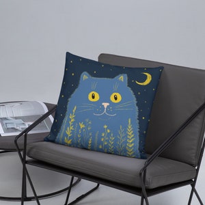 May include: A decorative square pillow featuring a whimsical illustration of a blue cat with yellow eyes, a crescent moon, and stars on a dark blue background. The cat is surrounded by yellow floral elements. The pillow is placed on a gray chair.