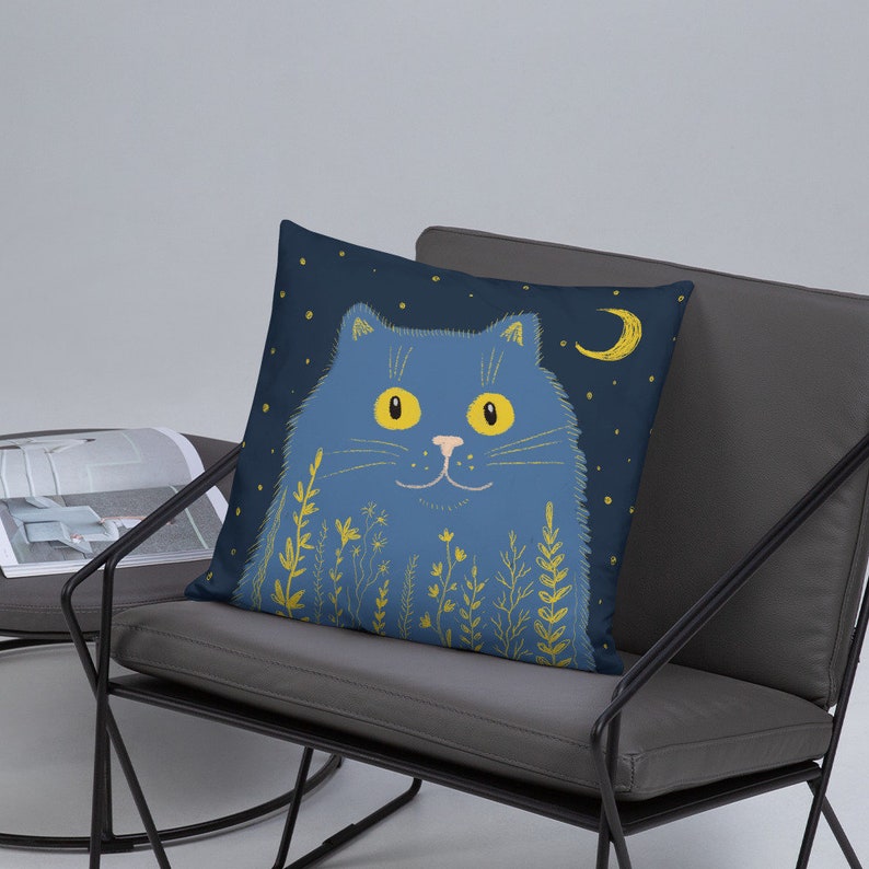 May include: A decorative square pillow featuring a whimsical illustration of a blue cat with yellow eyes, a crescent moon, and stars on a dark blue background. The cat is surrounded by yellow floral elements. The pillow is placed on a gray chair.