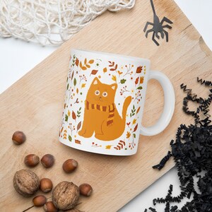 May include: White ceramic mug with a cartoon orange cat wearing a brown and yellow scarf. The cat is surrounded by a pattern of fall leaves and flowers in shades of orange, yellow, brown, and green.