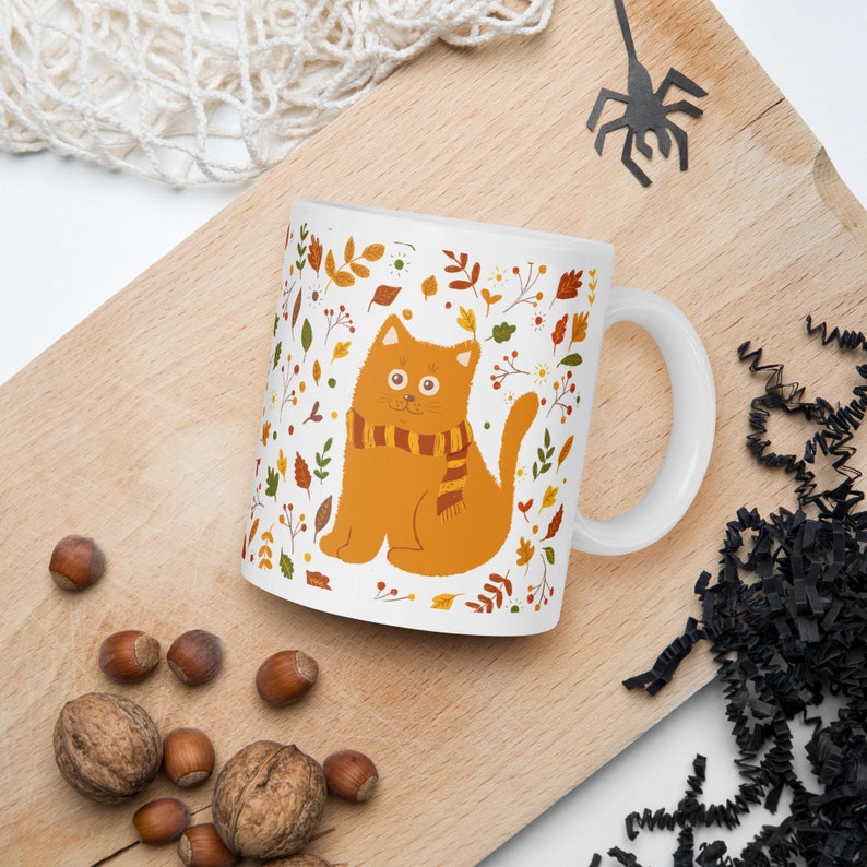 May include: White ceramic mug with a cartoon orange cat wearing a brown and yellow scarf. The cat is surrounded by a pattern of fall leaves and flowers in shades of orange, yellow, brown, and green.