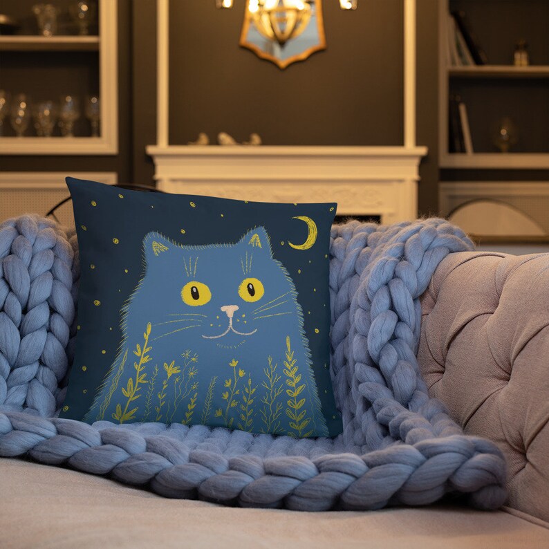 May include: A square throw pillow featuring a blue cat illustration against a dark blue background with a crescent moon and stars. The cat has yellow eyes and is surrounded by yellow floral accents. The pillow is on a light blue knitted blanket.