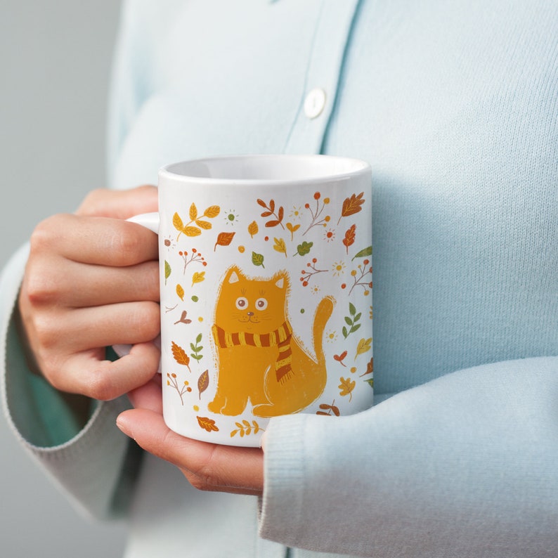 May include: White ceramic mug with a colorful autumn-themed design featuring a cat wearing a scarf. The design includes leaves, flowers, and berries.