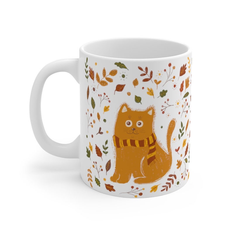 May include: White ceramic mug with a brown cat wearing a yellow and orange scarf. The cat is surrounded by a pattern of leaves and flowers in shades of brown, yellow, orange, and green.