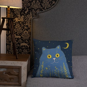 May include: A square decorative pillow featuring a blue cat illustration against a dark blue background with a crescent moon and stars. The cat has yellow eyes and is surrounded by yellow floral accents. The pillow is on a gray bed.