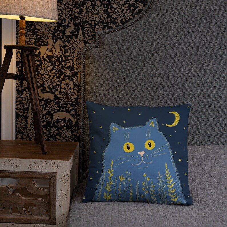 May include: A square decorative pillow featuring a blue cat illustration against a dark blue background with a crescent moon and stars. The cat has yellow eyes and is surrounded by yellow floral accents. The pillow is on a gray bed.