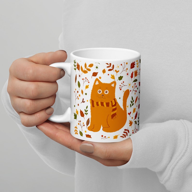 May include: White ceramic mug with a colorful autumn-themed design featuring a cat wearing a scarf. The cat is orange and the design includes leaves, flowers, and berries.