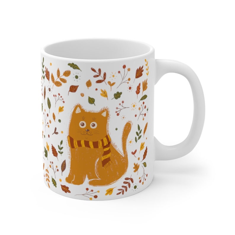 May include: White ceramic mug with a brown cat wearing a striped scarf. The cat is surrounded by a pattern of autumn leaves and flowers in shades of orange, yellow, and brown.