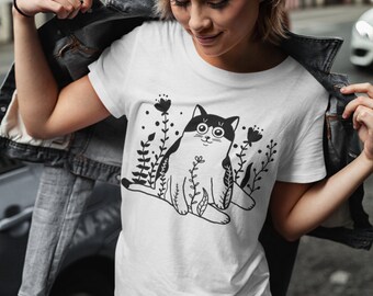 May include: White t-shirt with a black and white illustration of a cat sitting in a field of flowers.