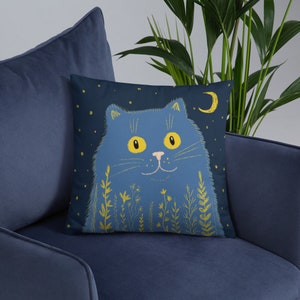 May include: A navy blue throw pillow featuring a whimsical illustration of a blue cat with yellow eyes, whiskers, and a pink nose. The cat is surrounded by yellow floral elements, stars, and a crescent moon on a dark blue background.