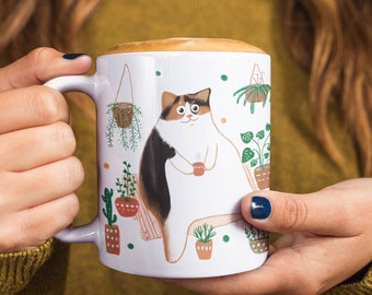 May include: White ceramic mug featuring a cartoon cat holding a small red cup, surrounded by potted plants and hanging planters. The cat has brown, black, and white fur. The mug is held by a person with a green sweater and dark blue nail polish.