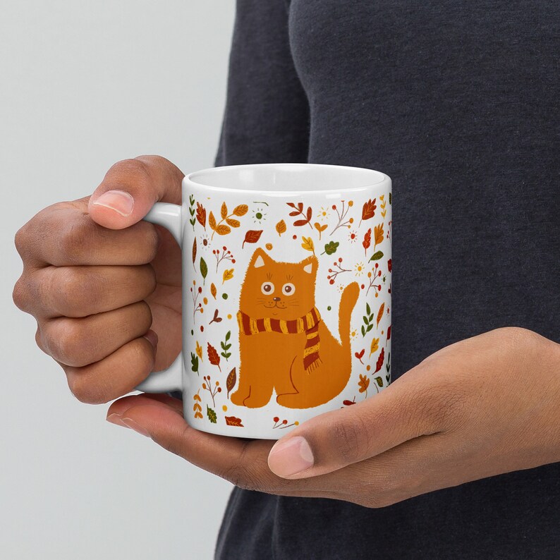 May include: White ceramic mug with a colorful autumn leaf design and a cartoon orange cat wearing a scarf.