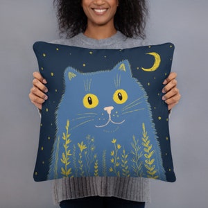 May include: A decorative pillow featuring a blue cat illustration against a dark blue background with a crescent moon and stars. The cat has yellow eyes and is surrounded by yellow floral elements.