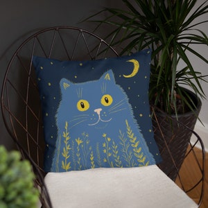 May include: A square throw pillow featuring a blue cat illustration against a dark blue starry night sky. The cat has yellow eyes and is surrounded by yellow floral accents. A yellow crescent moon is also visible.