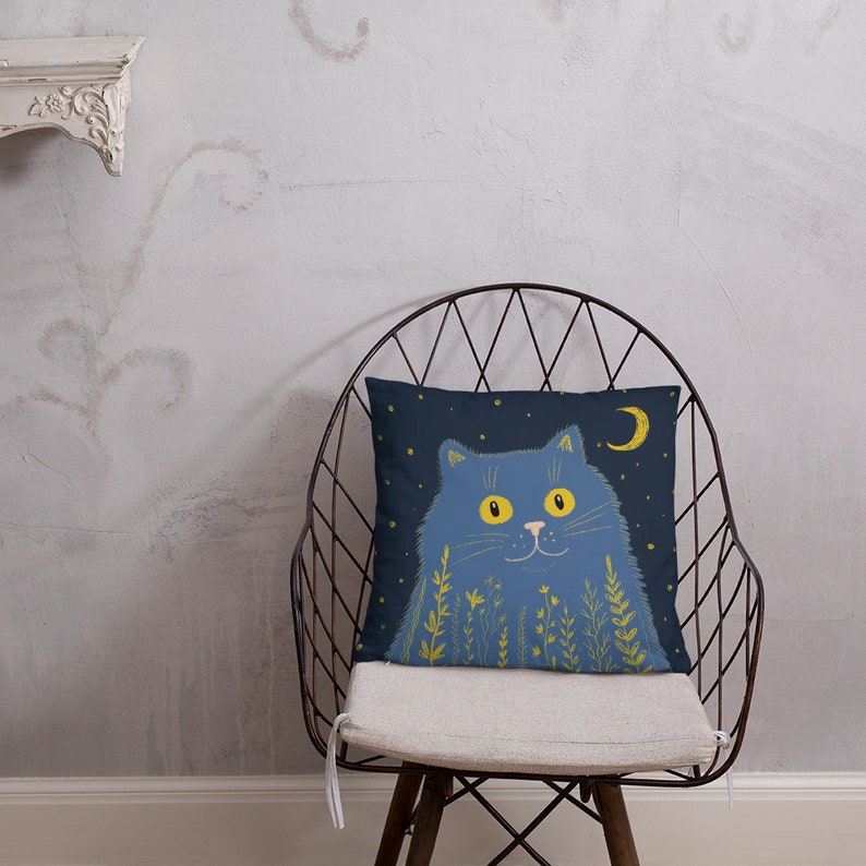 May include: A decorative throw pillow featuring a blue cat illustration against a dark blue background with a crescent moon and stars. The cat has yellow eyes and is surrounded by yellow floral accents. The pillow is on a metal chair.