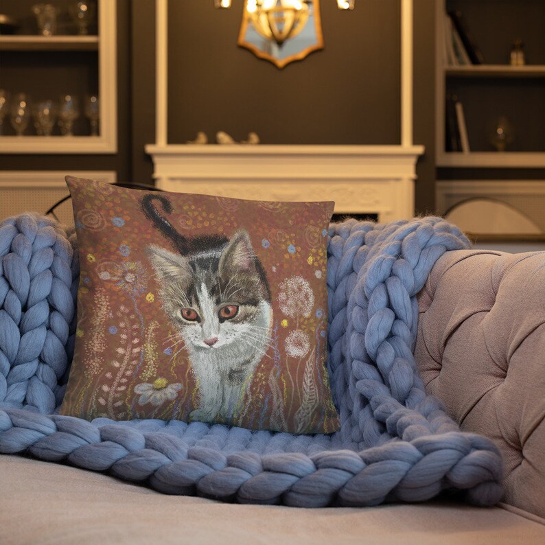 May include: A throw pillow with a brown background featuring a painting of a tabby cat with white markings. The cat is looking directly at the viewer. The background is filled with a variety of flowers and plants in shades of pink, yellow, and green.