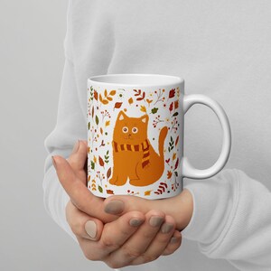 May include: White ceramic mug with a colorful autumn-themed design featuring a cat wearing a scarf. The cat is orange and the design includes leaves, flowers, and berries.