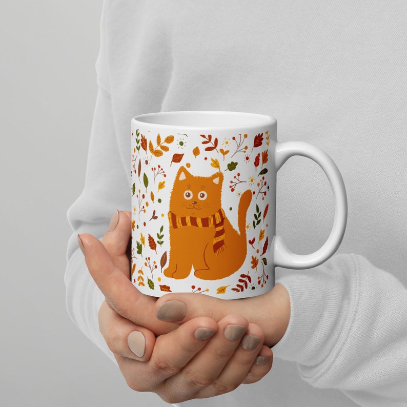 May include: White ceramic mug with a colorful autumn-themed design featuring a cat wearing a scarf. The cat is orange and the design includes leaves, flowers, and berries.