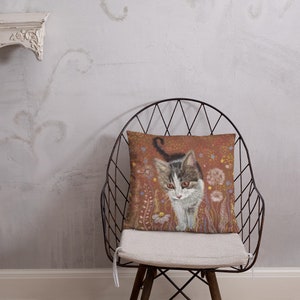 May include: A throw pillow with a brown background featuring a white and gray kitten walking through a field of flowers. The flowers are in shades of pink, yellow, and orange.